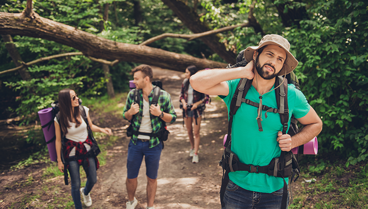 A picture of a man on a hike with his friends swatting at a mosquito on his neck.